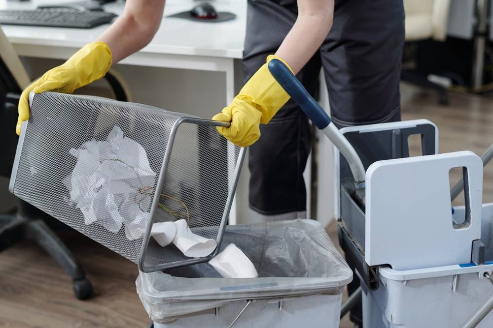Reinigungskraft in gelben Handschuhen leert einen Papierkorb unter einem Schreibtisch