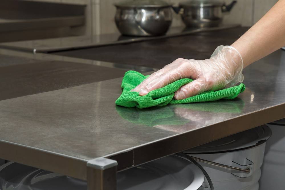 Mitarbeiterin mit Handschuh wischt eine Edelstahl-Arbeitsfläche in einer Großküche mit einem grünen Mikrofasertuch