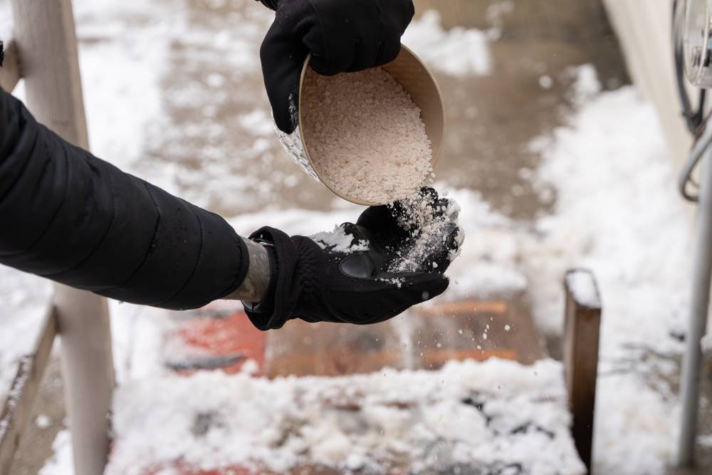 Hand im Handschuh streut Streugut auf einen verschneiten Weg